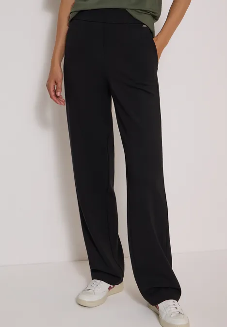 Une personne porte un pantalon noir ample et des baskets blanches, debout sur fond clair.