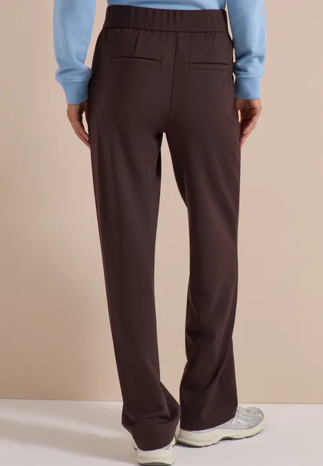 Vue de dos d'une personne en pantalon marron foncé ample et baskets blanches devant un fond beige.