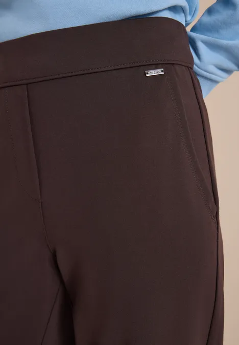 Gros plan sur un pantalon marron foncé élégant avec un petit logo argenté à la taille, porté avec un haut bleu.