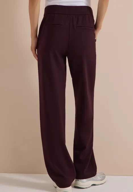 Vue de dos d’une personne portant un pantalon large bordeaux à taille élastique et des baskets claires sur fond beige.