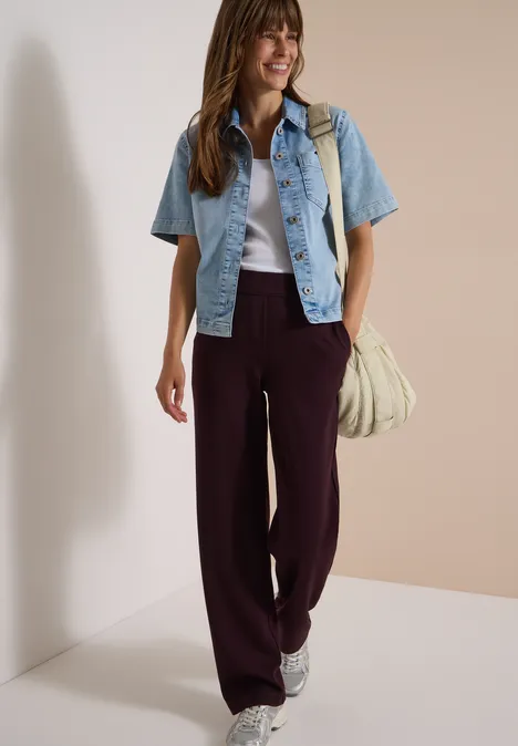 Femme souriante en pantalon bordeaux large, top blanc, veste courte en jean et baskets.