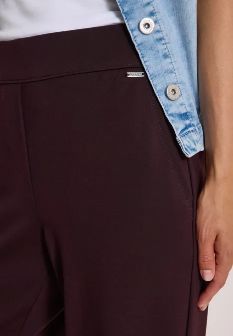Gros plan sur une personne en pantalon bordeaux, haut blanc et veste en jean, main sur la cuisse