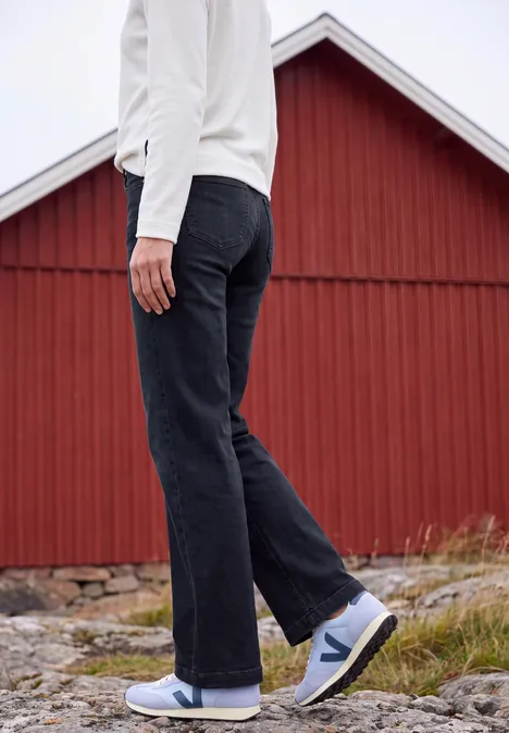 High Waist Wide Leg Jeans w kroju Loose Fit blue black used wash Osoba w białej kurtce, ciemnych dżinsach i białych butach przed czerwonym drewnianym budynkiem.