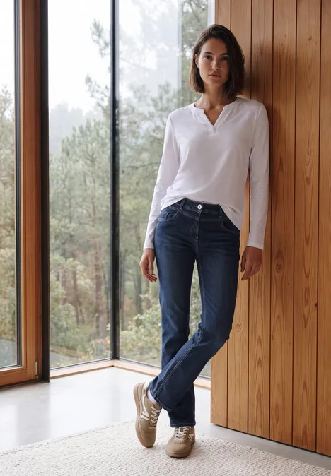 Eine Frau steht lässig in Jeans und weißer Bluse an einer Holzwand neben einem Fenster mit Waldblick.