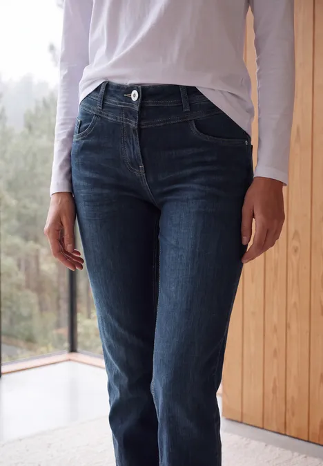 Eine Person in dunkelblauen Jeans und weißem Oberteil steht vor einem Fenster neben einer Holzverkleidung.