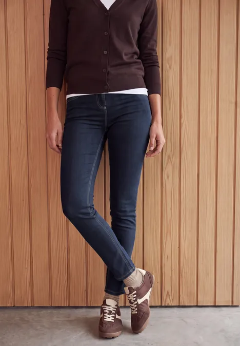 Unterkörper einer Person in dunklen Skinny-Jeans, braunen Sneakern und Cardigan vor Holzverkleidung.