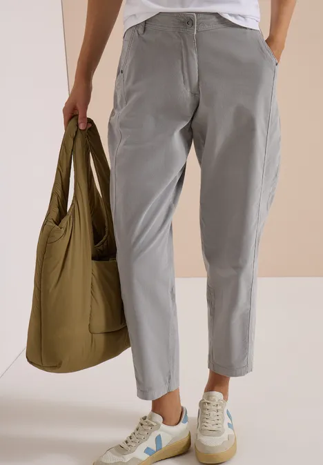Person trägt graue, lockere Hose, weiße Sneakers und hält eine olivgrüne Umhängetasche.