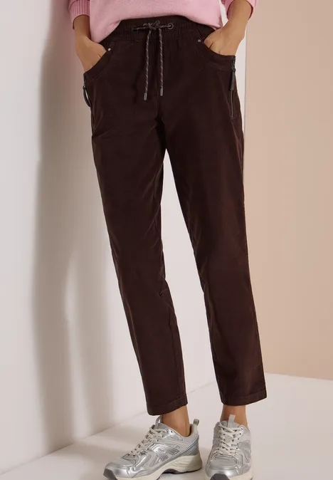 Mid Waist Slim Leg Hose im Casual Fit truffle brown Dunkelbraune Cordhose mit Kordelzug und lässigem geradem Schnitt, kombiniert mit silbernen Sneakers vor beiger Wand.