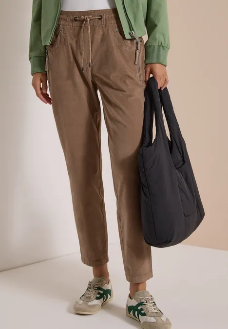 Mid Waist Slim Leg Hose im Casual Fit earthy taupe Person trägt braune Cordhose, grüne Jacke, beige Sneaker mit grünen Akzenten und schwarzen Shopper.