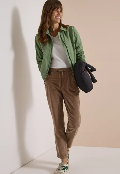Mid Waist Slim Leg Hose im Casual Fit earthy taupe Eine lächelnde Frau lehnt an der Wand, trägt grüne Jacke, weißes Shirt, braune Hose und hält eine gesteppte Tasche.