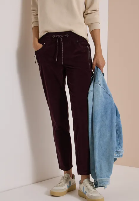 Mid Waist Slim Leg Hose im Casual Fit berry red Frau trägt burgundy Cordhose, cremefarbener Pullover, weiße Sneaker, hält hellblaue Jeansjacke.