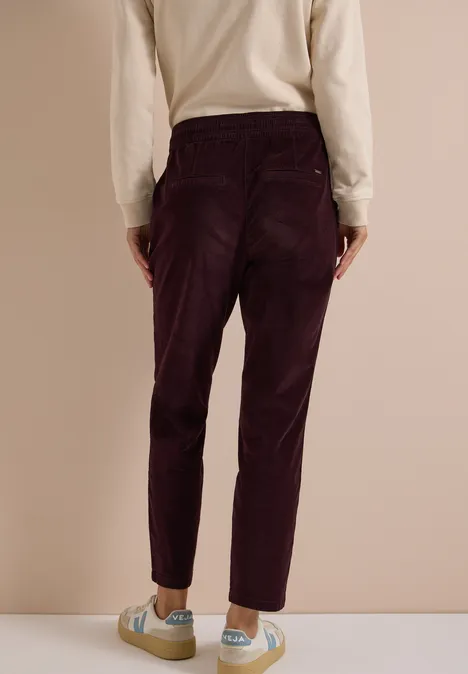 Mid Waist Slim Leg Hose im Casual Fit berry red Modell steht von hinten, trägt hochgeschnittene dunkelviolette Cordhose, beiges Oberteil und helle Sneaker.