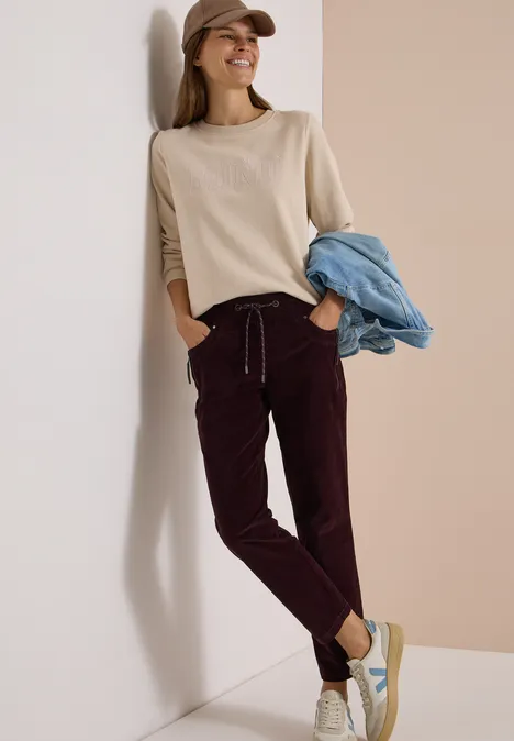 Mid Waist Slim Leg Hose im Casual Fit berry red Frau lehnt an Wand, trägt beige Pullover, weinrote Hose, Sneaker, hält blaue Jeansjacke.