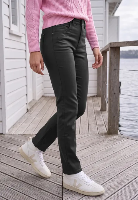Mid Waist Straight Leg Hose im Slim Fit Black Eine Person steht auf einem hölzernen Steg am Wasser, trägt schwarze Jeans, weiße Sneaker und einen rosafarbenen Pullover.