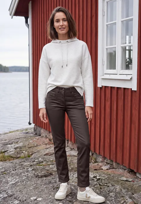 Eine Frau steht am Ufer neben einem roten Holzhaus am See, trägt weißen Hoodie, braune Hose und weiße Sneaker.