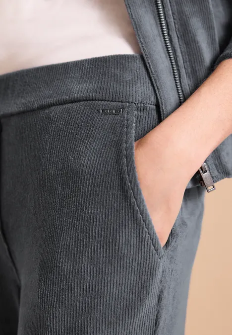 Spodnie z High Waist i prostymi nogawkami w stylu Loose Fit stone ash grey Zbliżenie na ciemnoszare sztruksowe spodnie z ręką w kieszeni i dopasowaną kurtką.