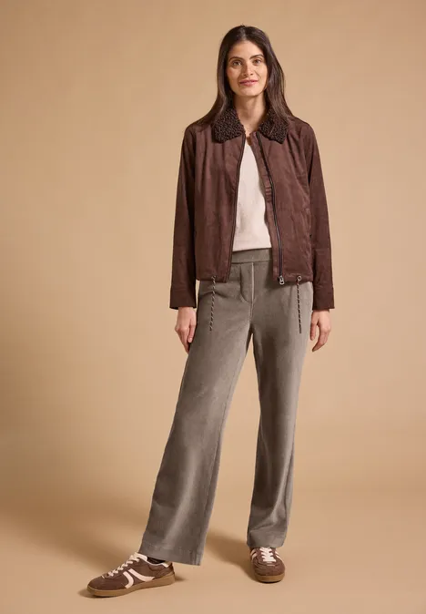 Spodnie z wysokim stanem i prostymi nogawkami w kroju Loose Fit earthy taupe Kobieta w brązowej zamszowej kurtce, białym topie, szarych szerokich spodniach i brązowych trampkach, na neutralnym tle.