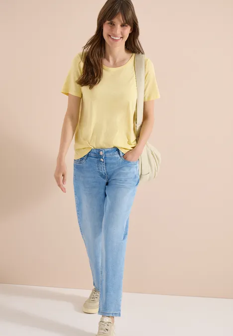 Een vrouw draagt een gele t-shirt, lichte spijkerbroek en sneakers, met een schoudertas tegen neutrale achtergrond.