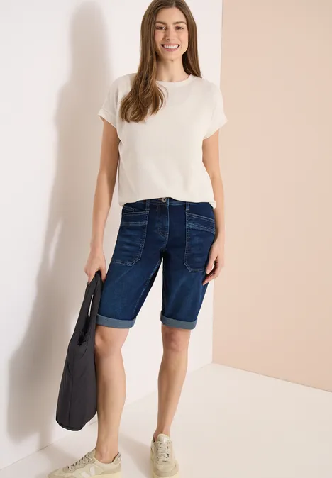 Frau in weißem T-Shirt und dunklen Bermudashorts, hält eine Tasche, trägt Sneaker.