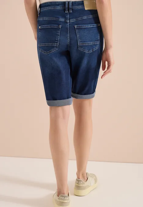 Rückseite einer Person in dunkelblauen Jeans-Bermuda-Shorts mit Umschlag und hellen Sneakern vor beiger Wand.