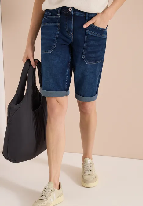 Person trägt dunkle Jeans-Shorts, weiße Schuhe und hält eine schwarze Stofftasche.