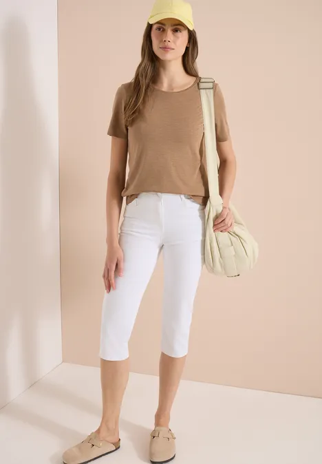 Une femme porte un t-shirt beige, un pantacourt blanc, des chaussures beiges et un sac assorti.