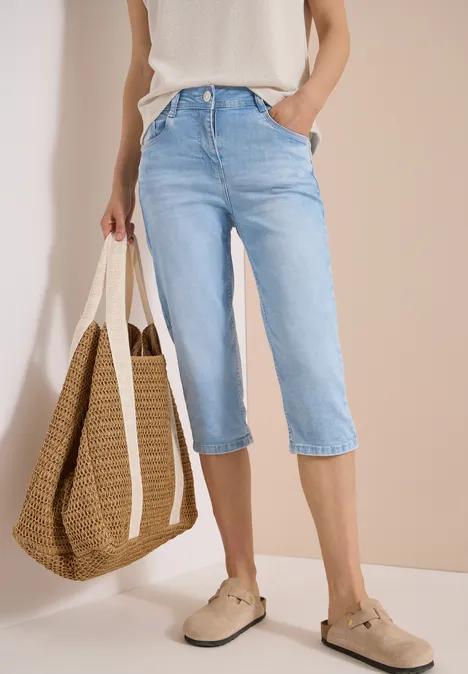 Frau in hellblauer Capri-Jeans mit weißem Oberteil, Basttasche und beigen Espadrilles vor neutralem Hintergrund.
