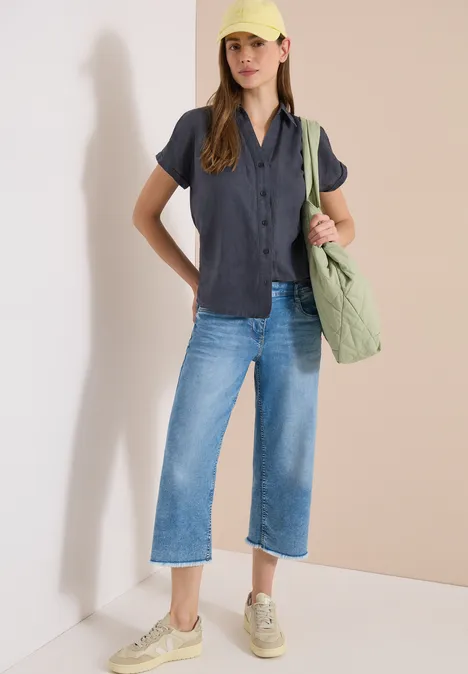 Vrouw leunt tegen muur, zwarte blouse met korte mouwen, wijde cropped jeans, beige sneakers en groene gewatteerde tas.