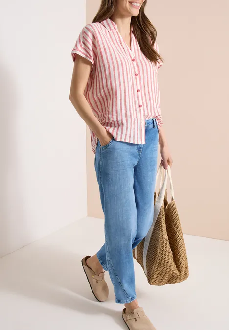 Dame met gestreept kort overhemd, blauwe jeans, beige schoenen en grote gevlochten tas.