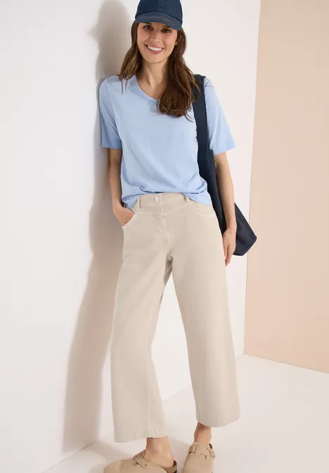 Une femme posée contre un mur clair porte un t-shirt bleu pâle, un pantalon large beige, une casquette et un sac en bandoulière.