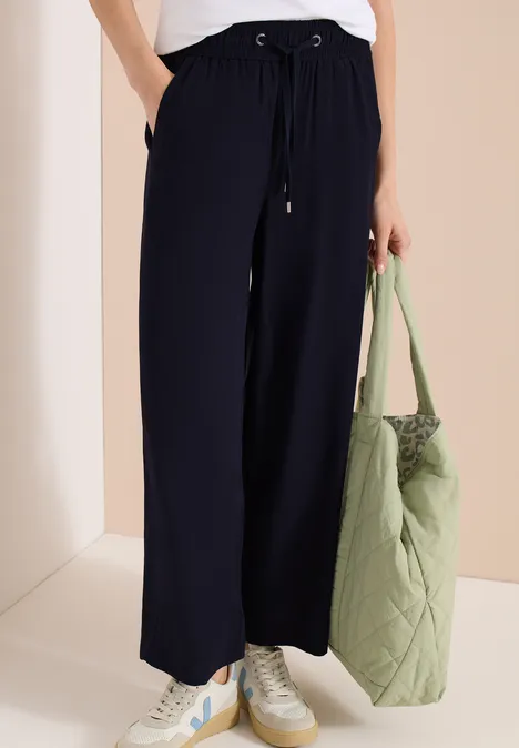 Frau trägt weite schwarze Hose mit Sneakers und hält eine große, gesteppt grüne Tasche.