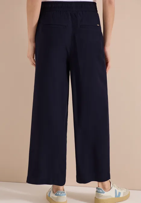 Rückenansicht einer Person in weiten, dunkelblauen Hosen und weißen Sneakern mit grünem Logo.