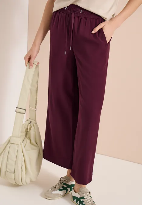 Frau trägt weinrote Jogginghose, weiße Sneaker und hält eine hellbeige Tasche vor neutralem Hintergrund.