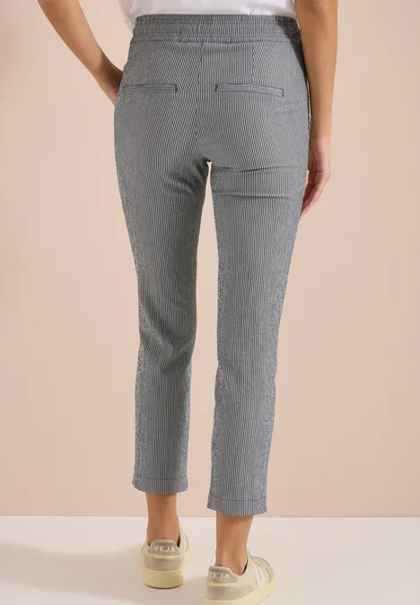Vrouw van achter in grijze, fijngestreepte 7/8-broek met elastische taille en sneakers, beige achtergrond.