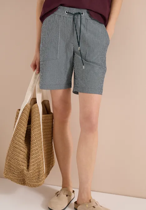 Person in gestreiften Shorts, trägt Strohtasche und bequeme Sandalen vor beigem Hintergrund.