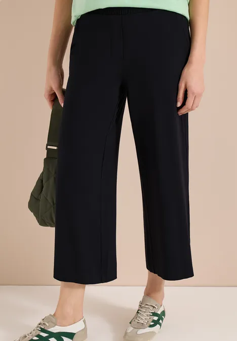 Frau trägt weite, knöchellange schwarze Hose, kombiniert mit weißen Sneakern und einer olivgrünen Tasche.