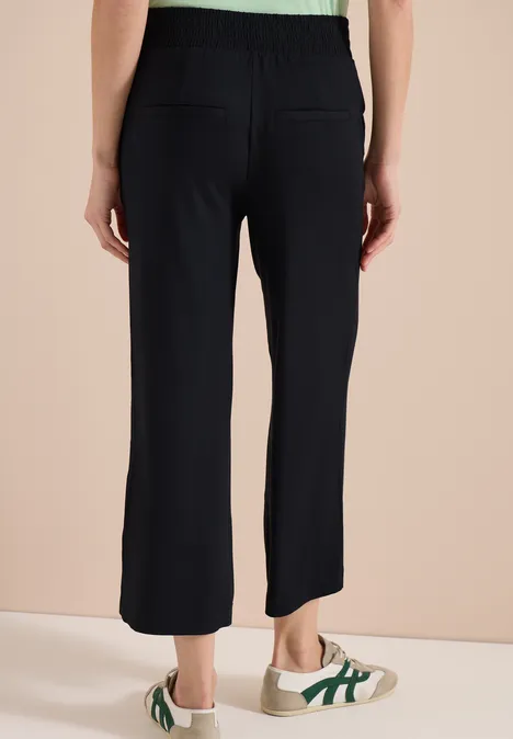 Rückenansicht einer Person in schwarzen, weiten Hosen und weißen Sneakern mit grünen Streifen vor beigem Hintergrund.