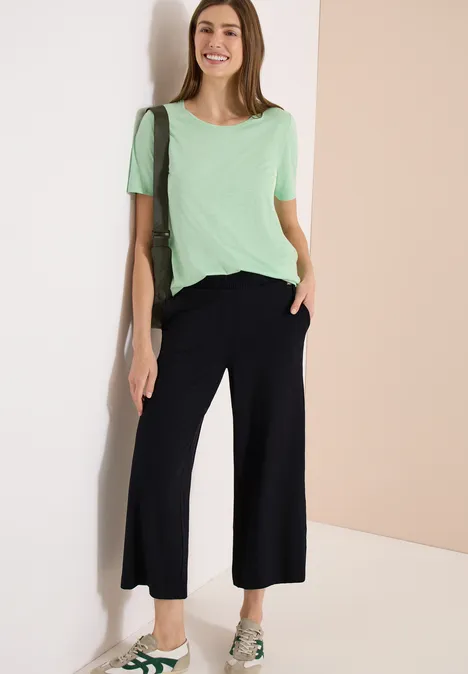 Frau lehnt an heller Wand, trägt mintgrünes T-Shirt, schwarze weite Culottes und Sandalen.