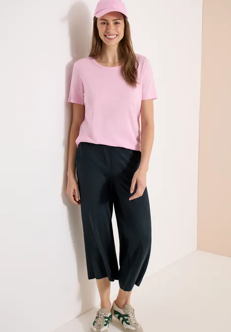 Frau in pinkem T-Shirt und weiten, schwarzen 3/4-Hosen steht vor heller Wand in einer Ecke.