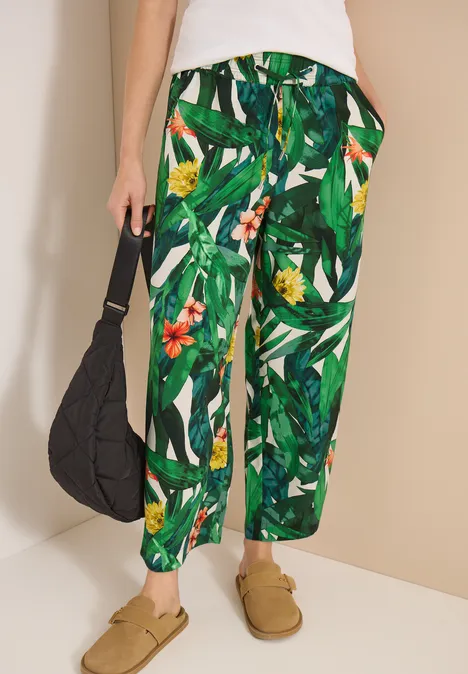 Grüne Stoffhose mit tropischem Blattmuster, lässiger Schnitt, schwarze Tasche und beige Slipper.