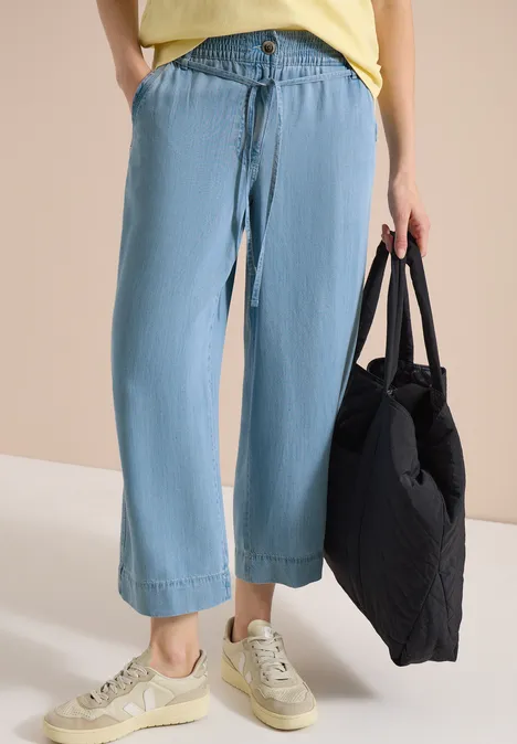 Iemand draagt wijde lichtblauwe jeans, beige sneakers en houdt een zwarte tas vast.