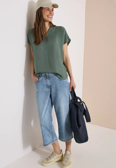 Frau in legerem Outfit: grünes T-Shirt, weite Jeanshose, Sneaker und Tasche lehnt entspannt an Wand.