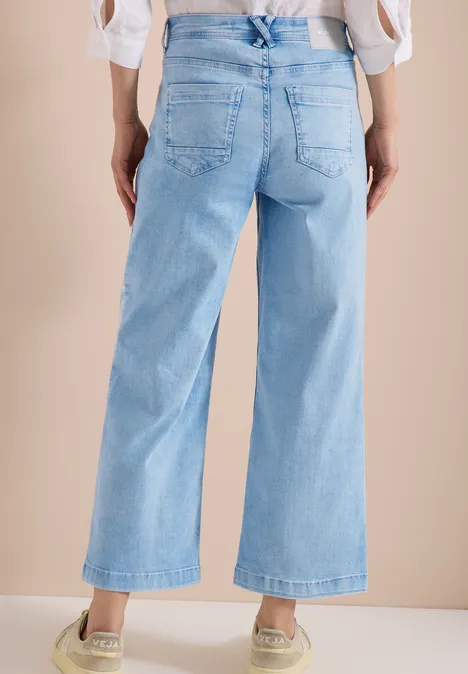 Rückansicht einer Person in hellblauen, weiten, knöchellangen Jeans mit Sneakers vor beigem Hintergrund.