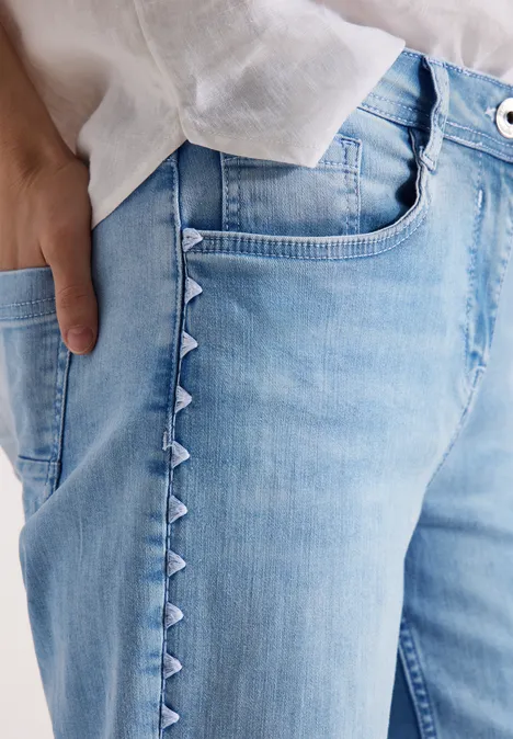 Helle Jeans mit seitlichen Dreieck-Details, weiße Bluse und Hand in Tasche.