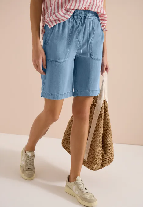 Jemand trägt blaue Jeans-Shorts, beige Sneaker, rosa-weiß gestreiftes Top und hält eine Strohtasche.