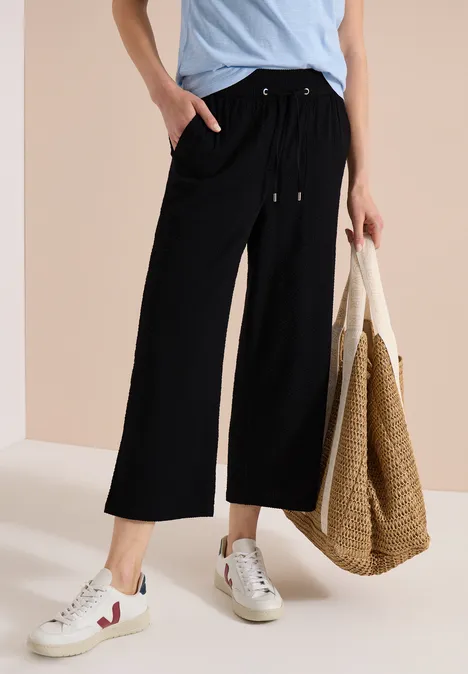 Zwarte culottebroek met trekkoord en brede pijpen, gecombineerd met sneakers en een rieten shopper.