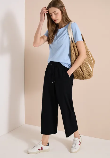 Vrouw met lichtblauw T-shirt, zwarte wijde cropped broek, witte sneakers en grote strozak op neutrale achtergrond.
