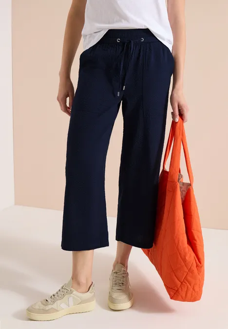 Vrouw met marineblauwe cropped broek, wit topje, beige sneakers en een oranje tas.