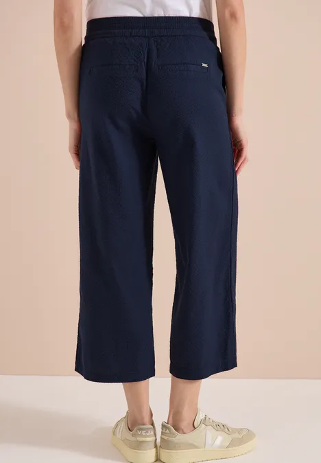 Achteraanzicht: wijde, donkerblauwe, enkelhoge broek met elastische taille, achterzak en lichte sneakers.