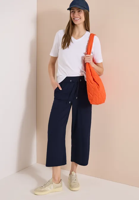 Vrouw met wit T-shirt, wijde donkerblauwe broek, beige sneakers, oranje tas en pet.